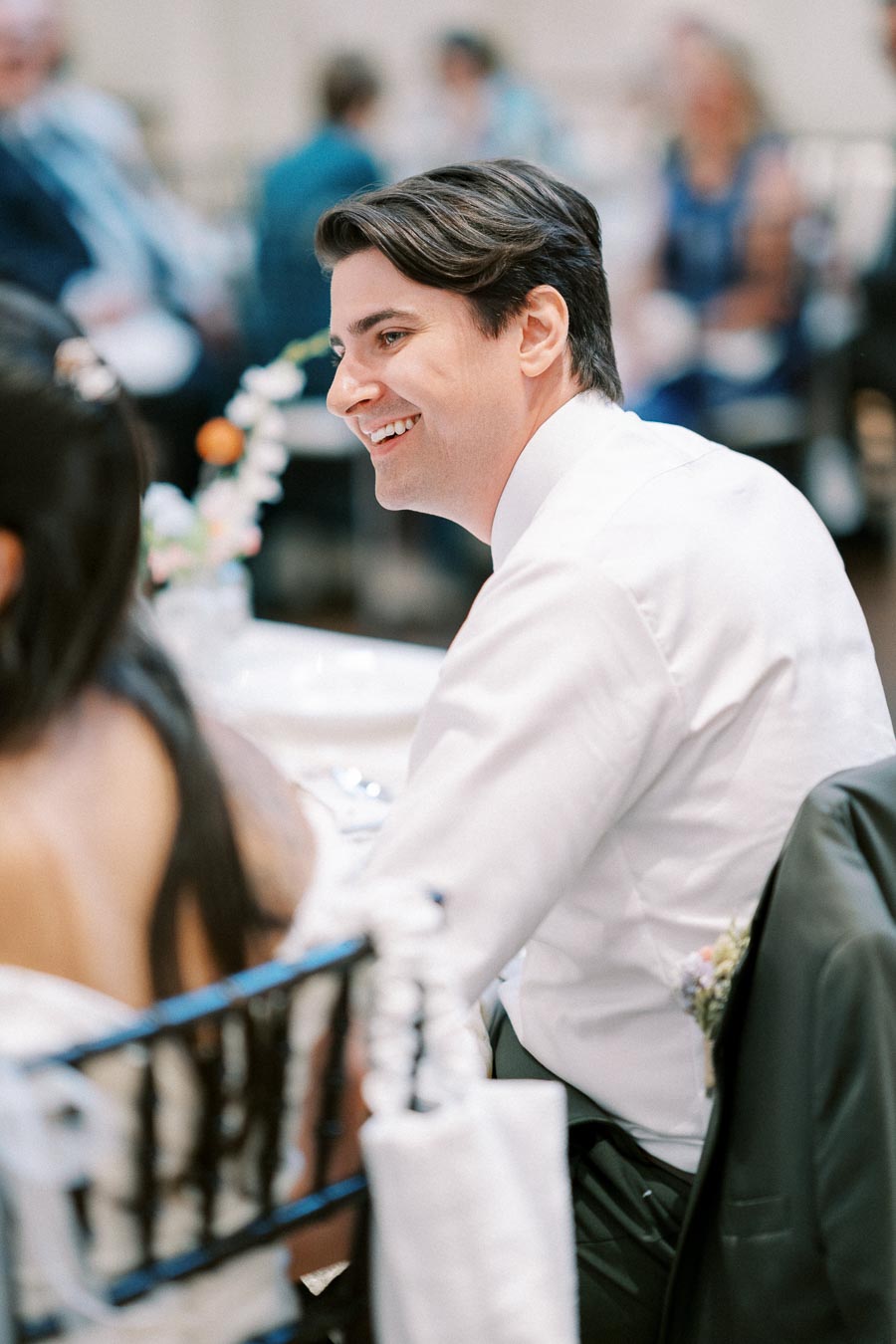 Smiling man in a white shirt at a formal event, sitting at a table decorated with flowers, in a blurred background of guests.
