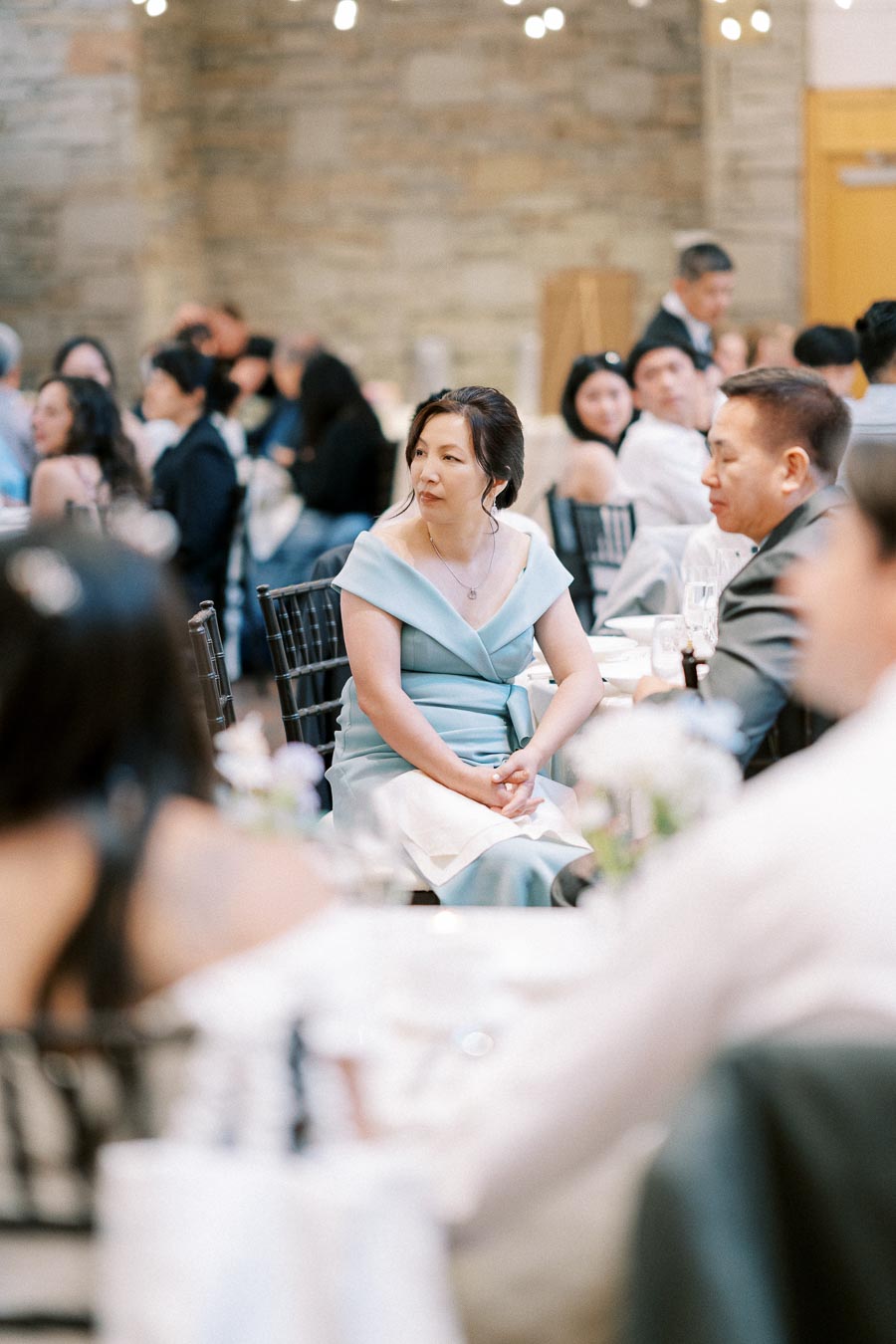 A woman in a light blue dress seated at a formal event, surrounded by other guests in a warmly lit setting.
