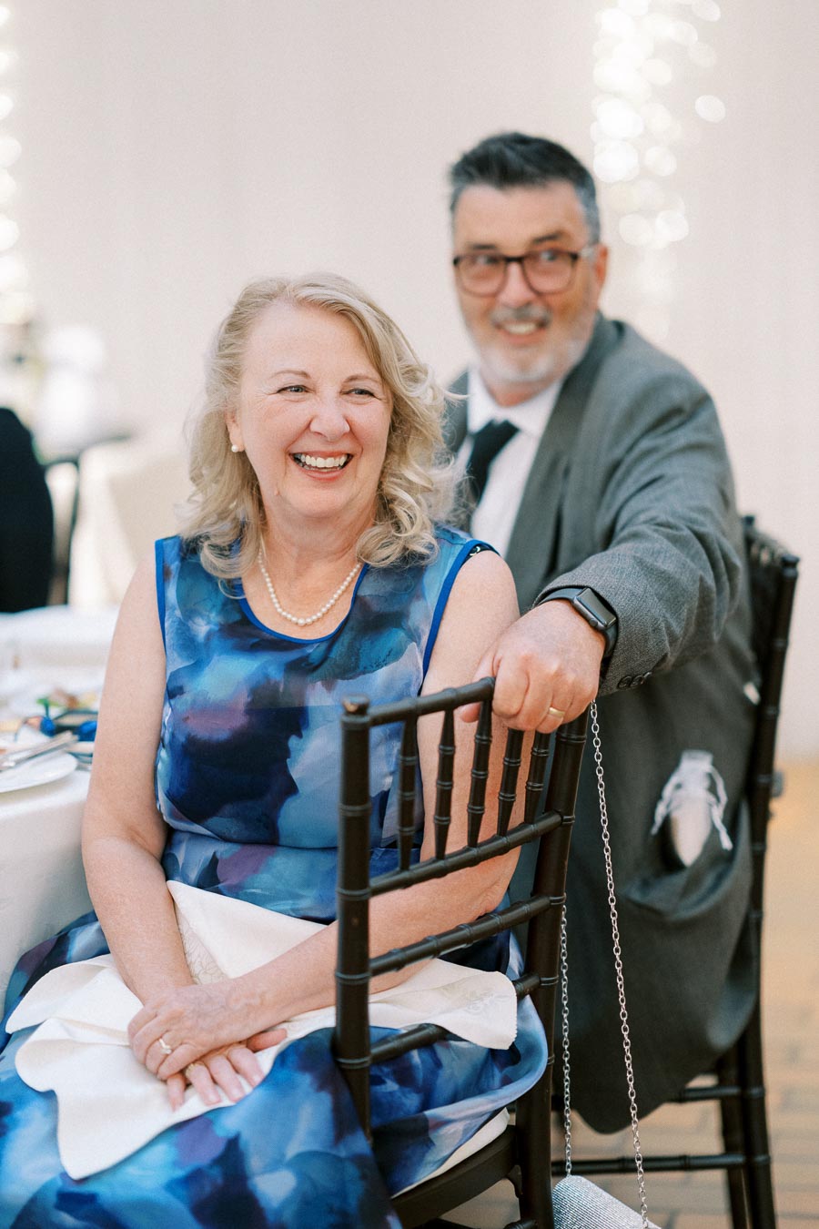 Senior couple smiling at a formal event, with the woman in a blue dress and pearls, and the man in a gray suit with glasses.