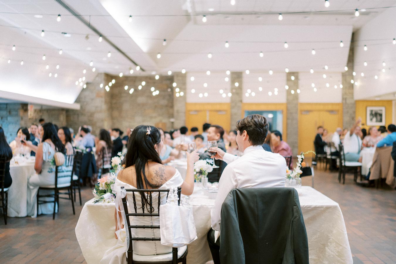 A couple toasting at a wedding reception in a beautifully decorated venue with string lights and elegantly adorned tables, surrounded by guests enjoying the celebration.