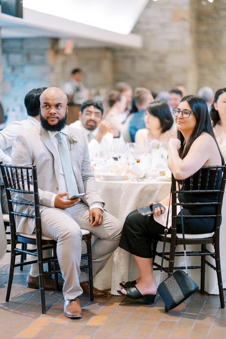 A group of people seated at a formal event, dressed in semi-formal attire, engaging in conversation around a table set with glassware and tableware, in a rustic stone venue.