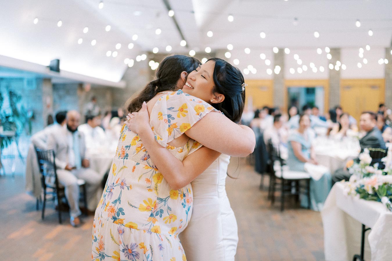 Two women embracing warmly at a wedding reception, adorned with string lights, surrounded by seated guests.