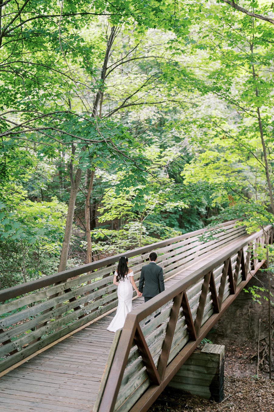A couple holding hands walks on a wooden bridge surrounded by lush green trees in a serene forest setting.