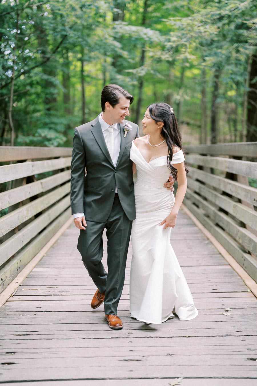 A bride and groom walking arm in arm across a wooden bridge in a lush, green forest. The groom is wearing a sharp, gray suit while the bride is dressed in an elegant white gown. They are gazing lovingly at each other, surrounded by nature's tranquil beauty.
