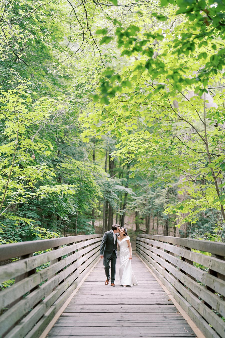 A couple in wedding attire walking on a wooden bridge surrounded by lush green trees in a forest setting.