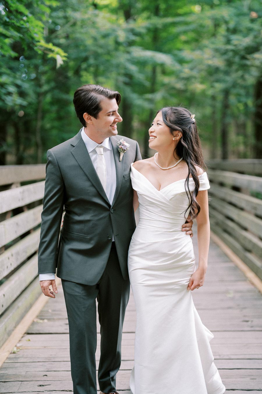 A joyful couple dressed in elegant wedding attire walking arm in arm on a wooden bridge surrounded by lush green trees. The groom is in a dark suit with a boutonniere, and the bride is wearing a white gown and pearl necklace.