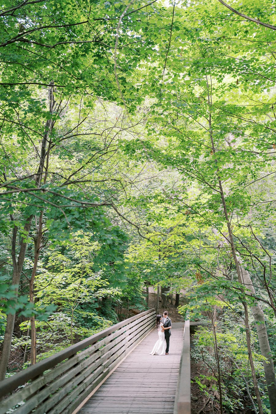 A couple embraces in wedding attire on a wooden bridge surrounded by lush green trees in a forest setting.