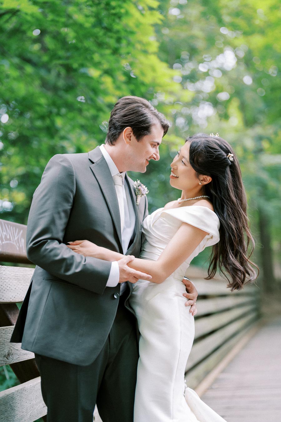A happy couple embracing on a wooden bridge surrounded by lush greenery, with the bride in an elegant white dress and the groom in a sharp suit, capturing a romantic moment in a natural setting.