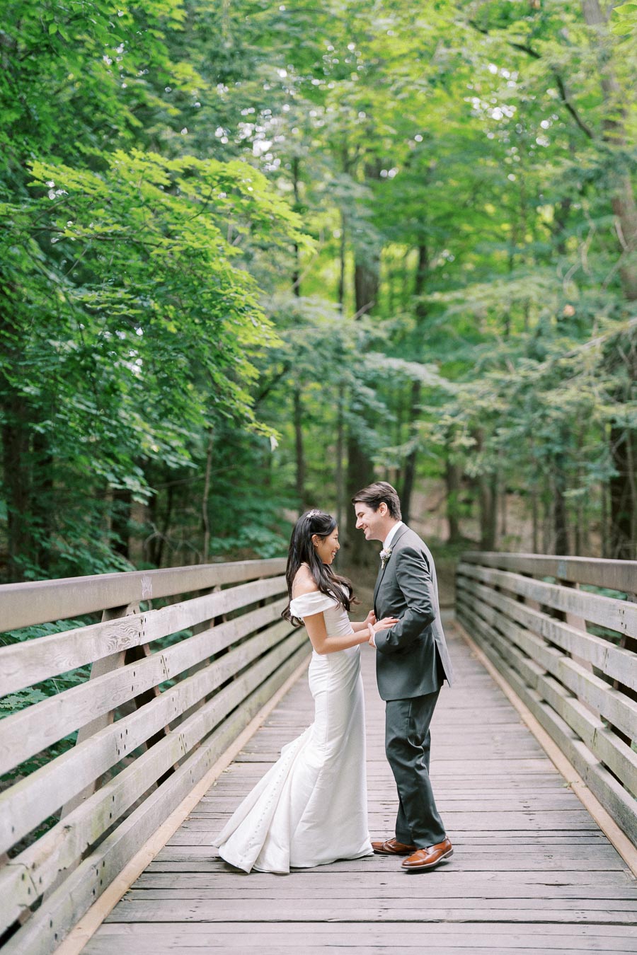 Bride and groom standing on a wooden bridge surrounded by lush green forest, sharing a romantic moment on their wedding day.