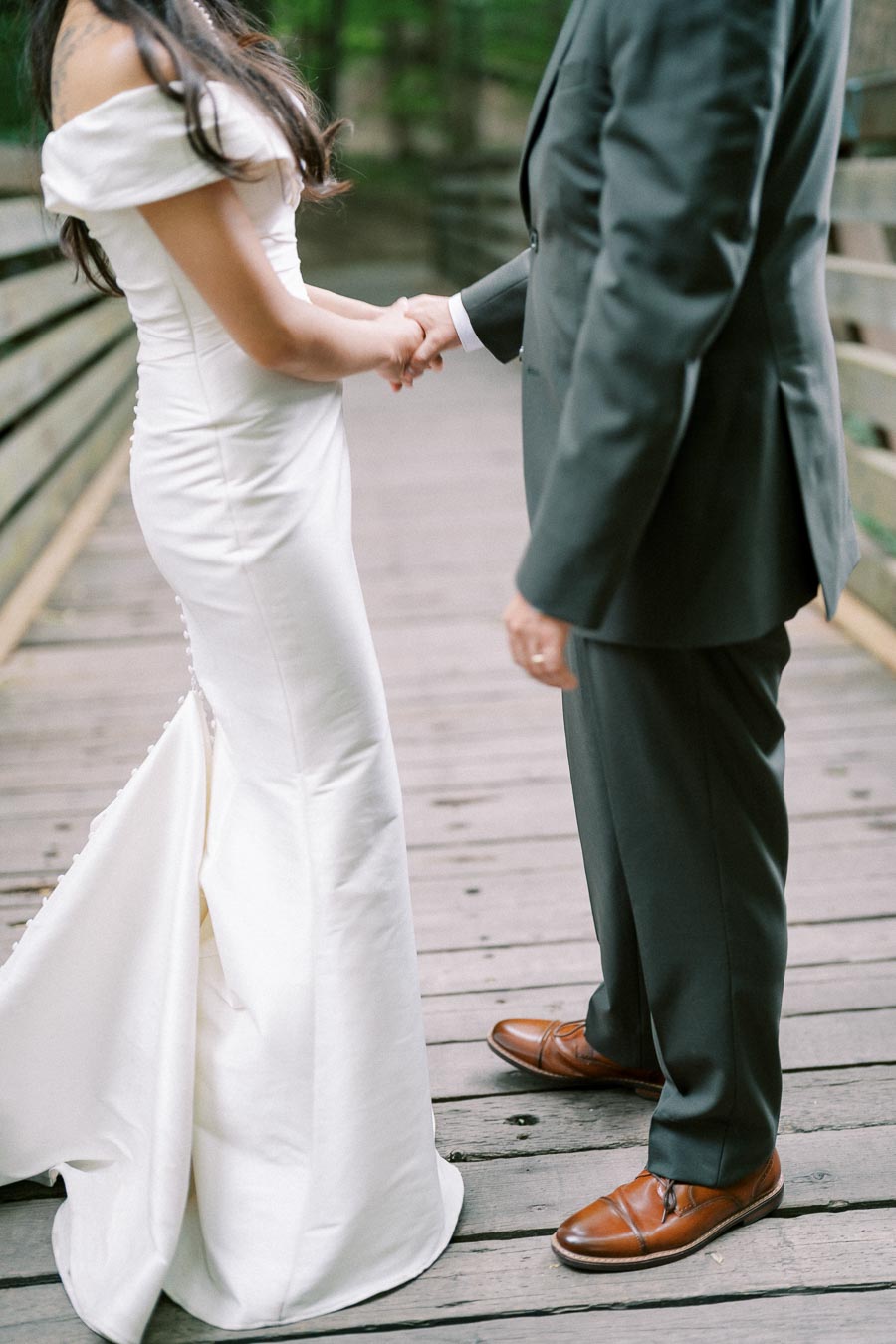 A bride in a white off-shoulder wedding dress and a groom in a dark suit holding hands on a wooden bridge surrounded by trees.