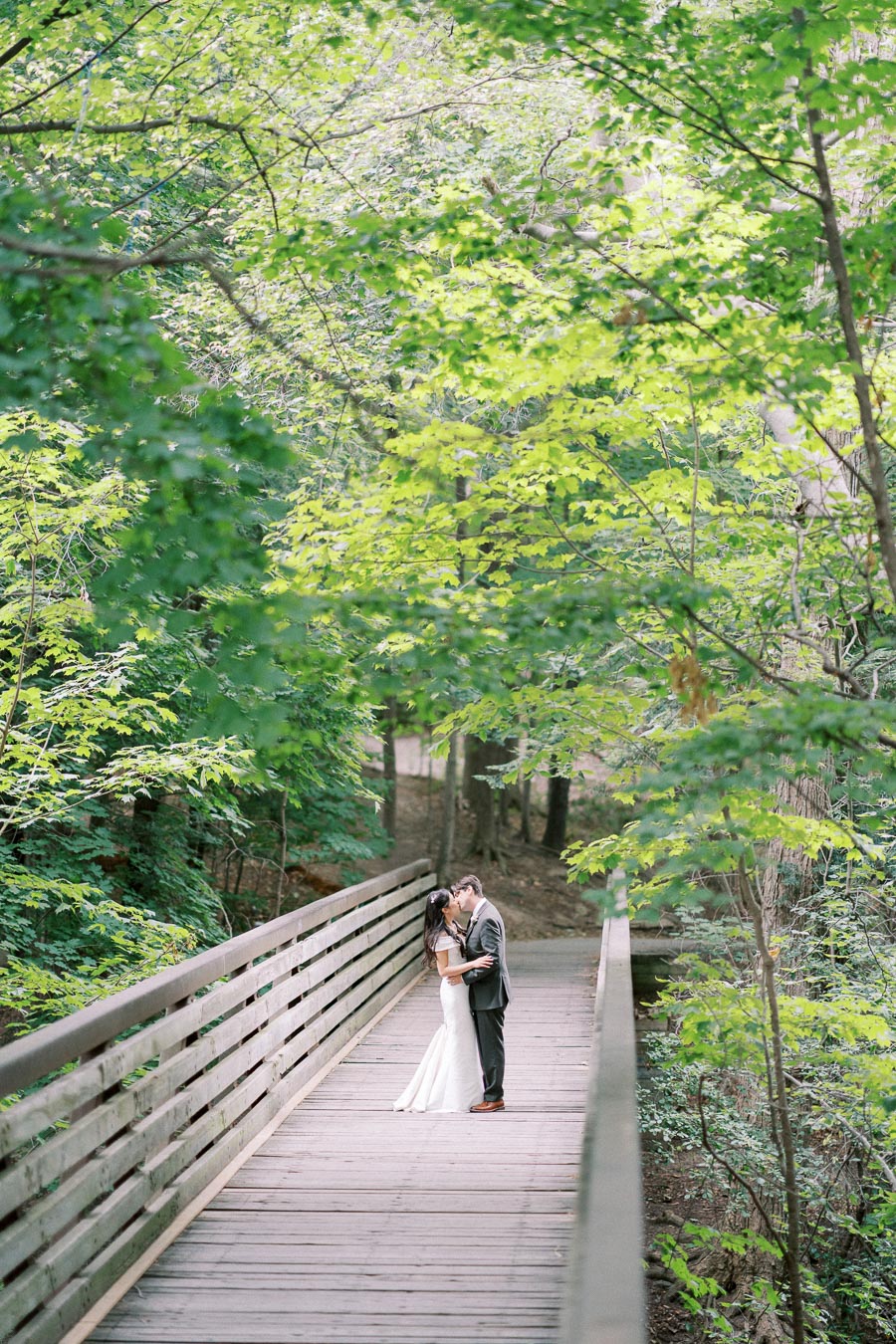 Romantic couple kissing on a wooden bridge surrounded by lush, green forest foliage.