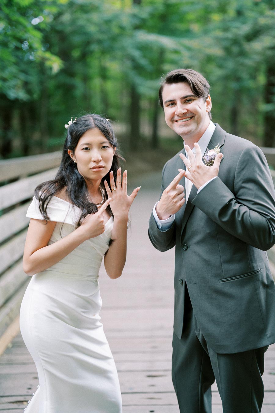 Bride and groom showing wedding rings while smiling on a picturesque wooden bridge with lush green trees in the background.