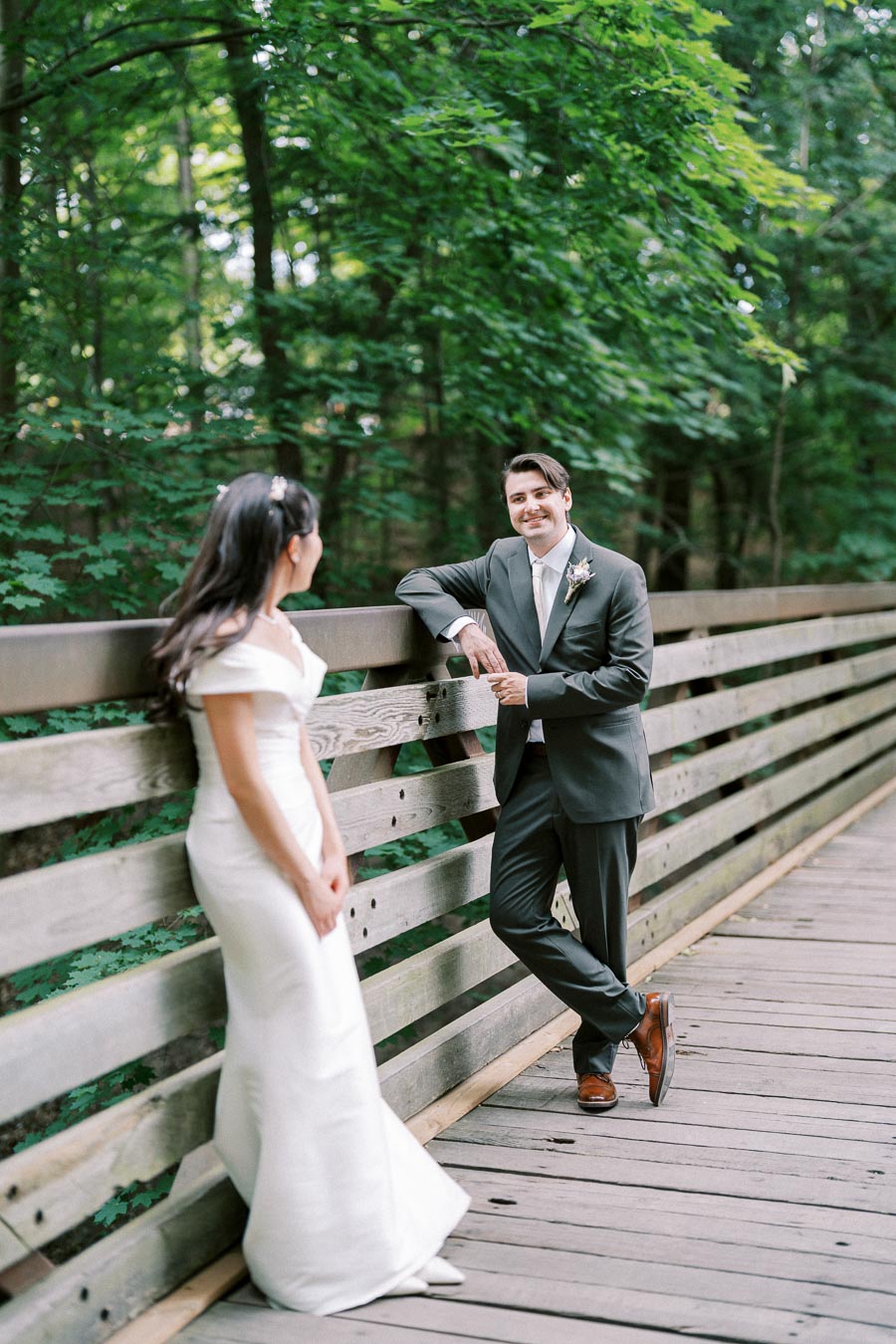 A bride in a white gown and a groom in a dark suit smile at each other on a wooden bridge surrounded by lush greenery, capturing a romantic wedding moment in nature.