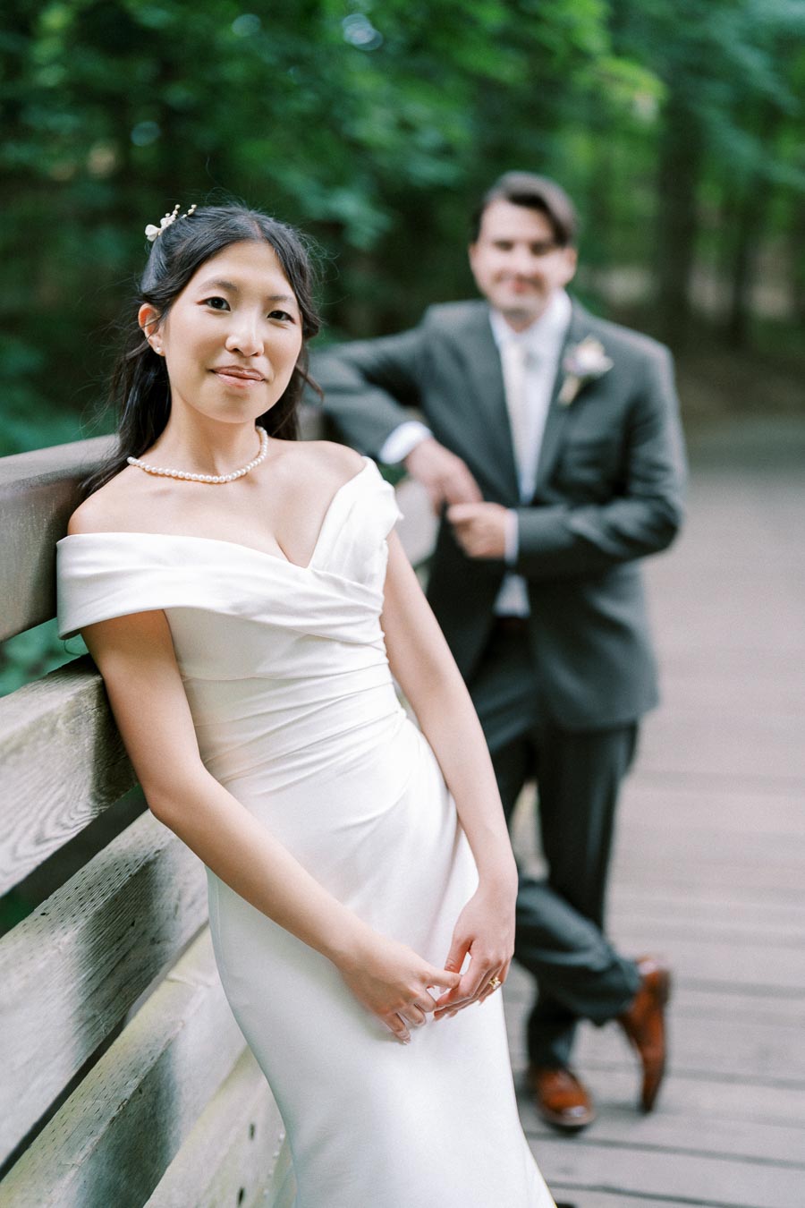 Bride in elegant white dress leans on wooden railing, with groom in a suit standing in the background, set in a lush green outdoor setting.