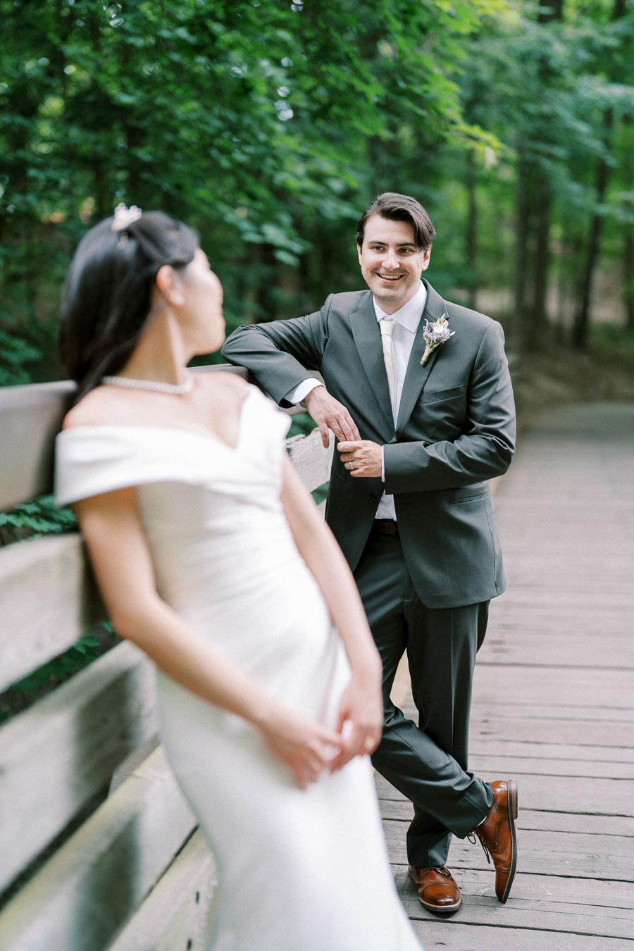 Bride in elegant white dress and groom in dark suit sharing a romantic moment on a wooden bridge, surrounded by lush green forest foliage.