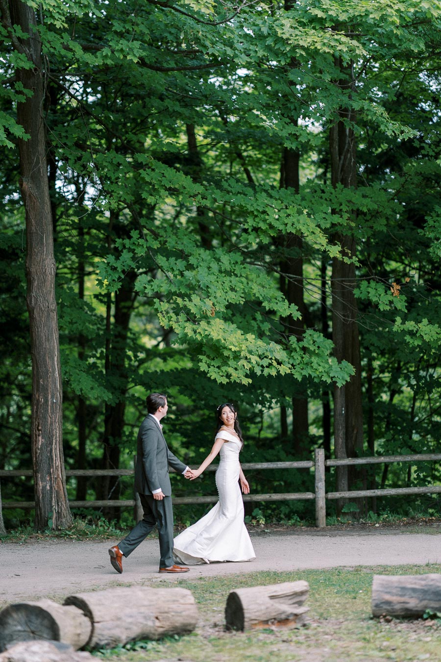 A bride and groom walk hand in hand along a rustic wooded path, surrounded by lush green trees. The bride wears a white gown, while the groom is in a dark suit, creating a picturesque wedding scene in nature.