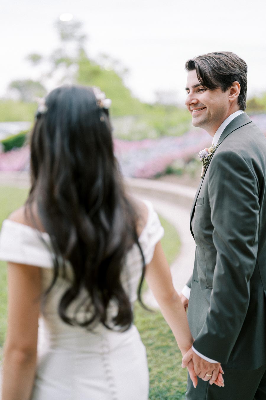 A bride and groom holding hands, smiling at each other, standing outdoors during their wedding ceremony in a lush garden setting.