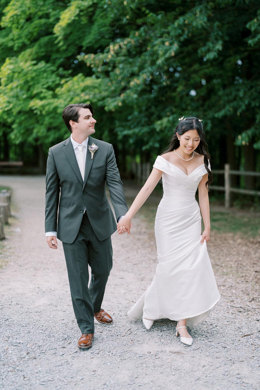 A newlywed couple, the groom in a dark suit and the bride in a white off-shoulder wedding gown, walking hand in hand on a tree-lined path.