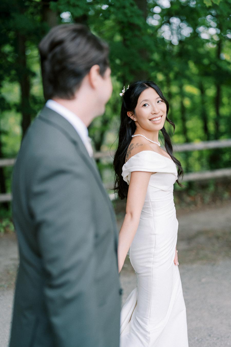 A bride in a white wedding dress smiles at her partner as they hold hands outdoors with green trees in the background.
