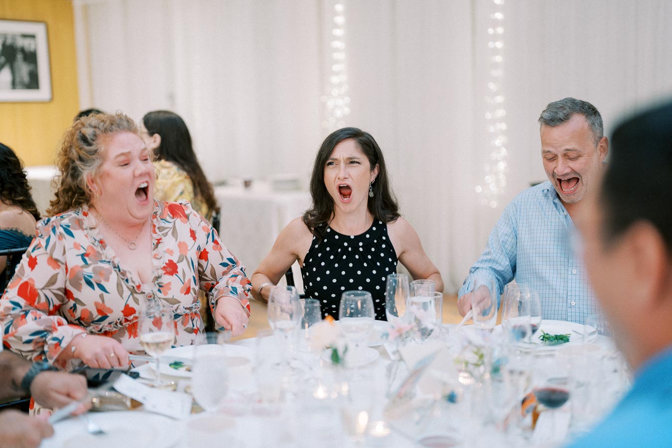 Group of people laughing and enjoying a meal together at a festive gathering, with drinks and decorations on a round table.