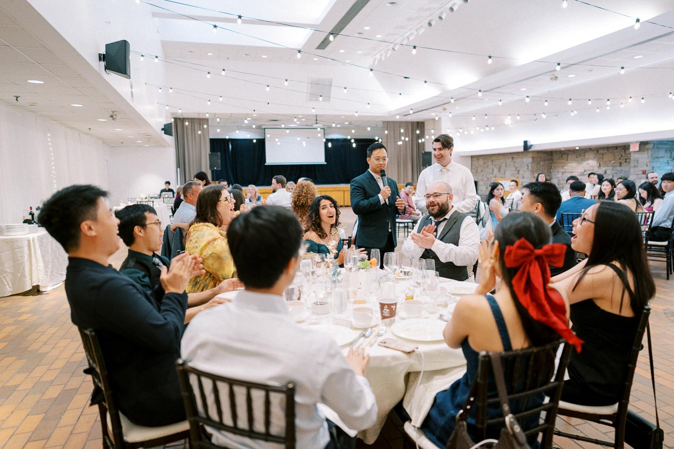 A group of people enjoying a formal banquet in a decorated hall, with a man speaking into a microphone while guests applaud at a table adorned with white tablecloths and various dishes.