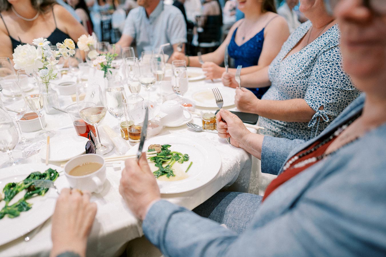 A group of people seated around an elegantly set table at a wedding reception, with plates of food, glasses of wine, and floral centerpieces, creating a festive and celebratory atmosphere.