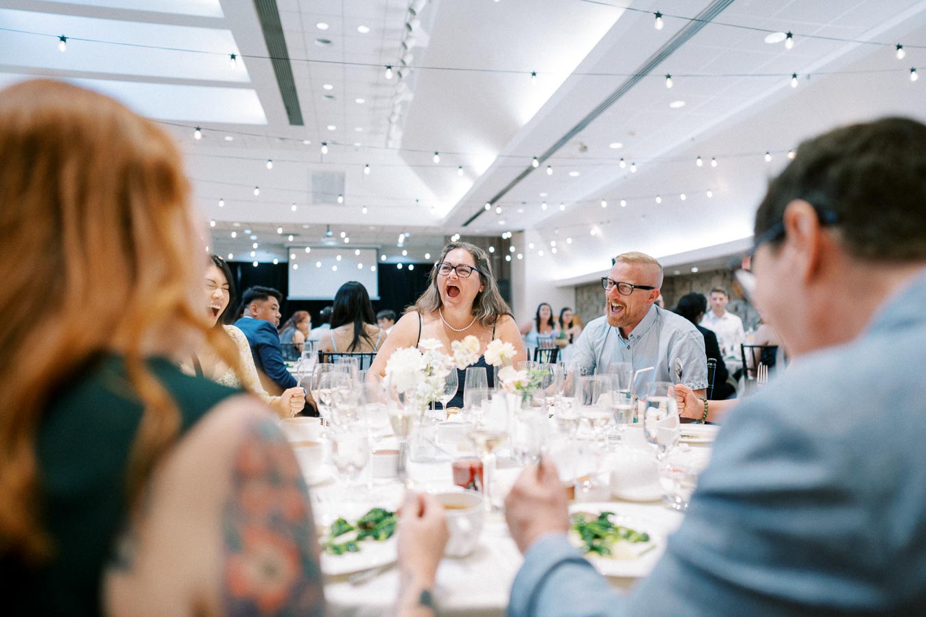 A group of people laughing and enjoying a meal together at a festive event, under a ceiling decorated with string lights.