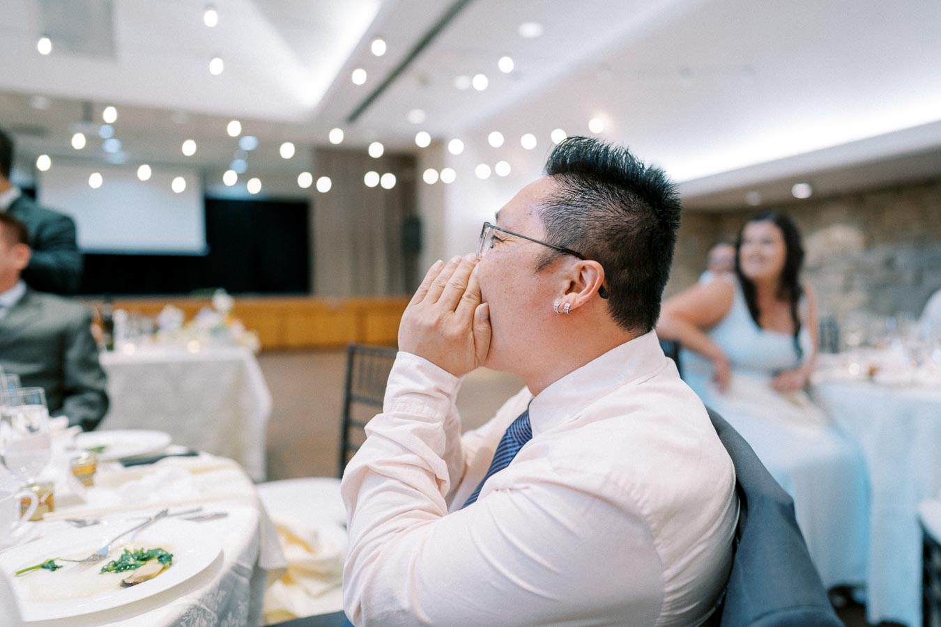 A person in a formal setting, seated at a round table with a white tablecloth, appearing engaged and excited, surrounded by elegantly dressed guests and soft ambient lighting.