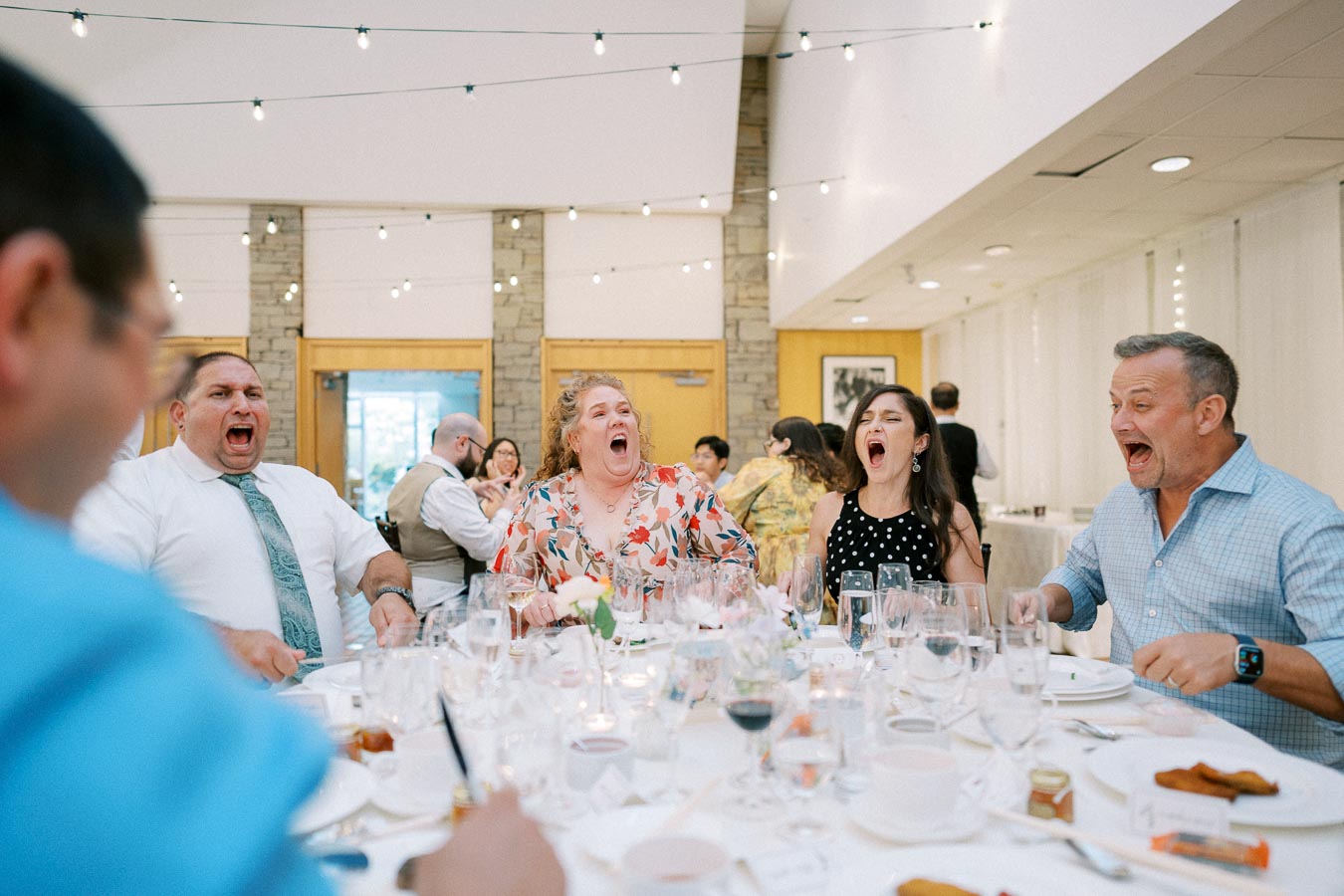 Group of people at a lively banquet table sharing laughter and joy during a social gathering, with glasses and plates on the table, under elegant string lights.