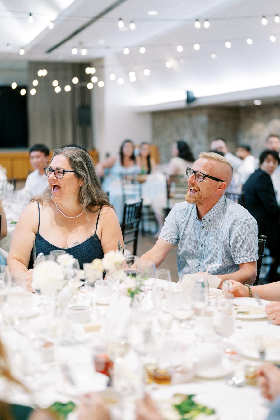 People laughing and enjoying a social event at a beautifully decorated venue with string lights and elegantly set tables.