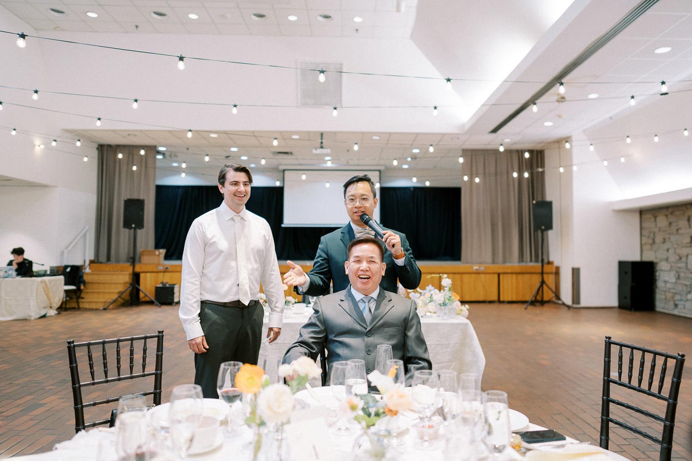 A joyful celebration in a beautifully decorated banquet hall featuring three men, with one seated and smiling widely, as another holds a microphone while speaking. The setting is adorned with string lights and elegant floral centerpieces.
