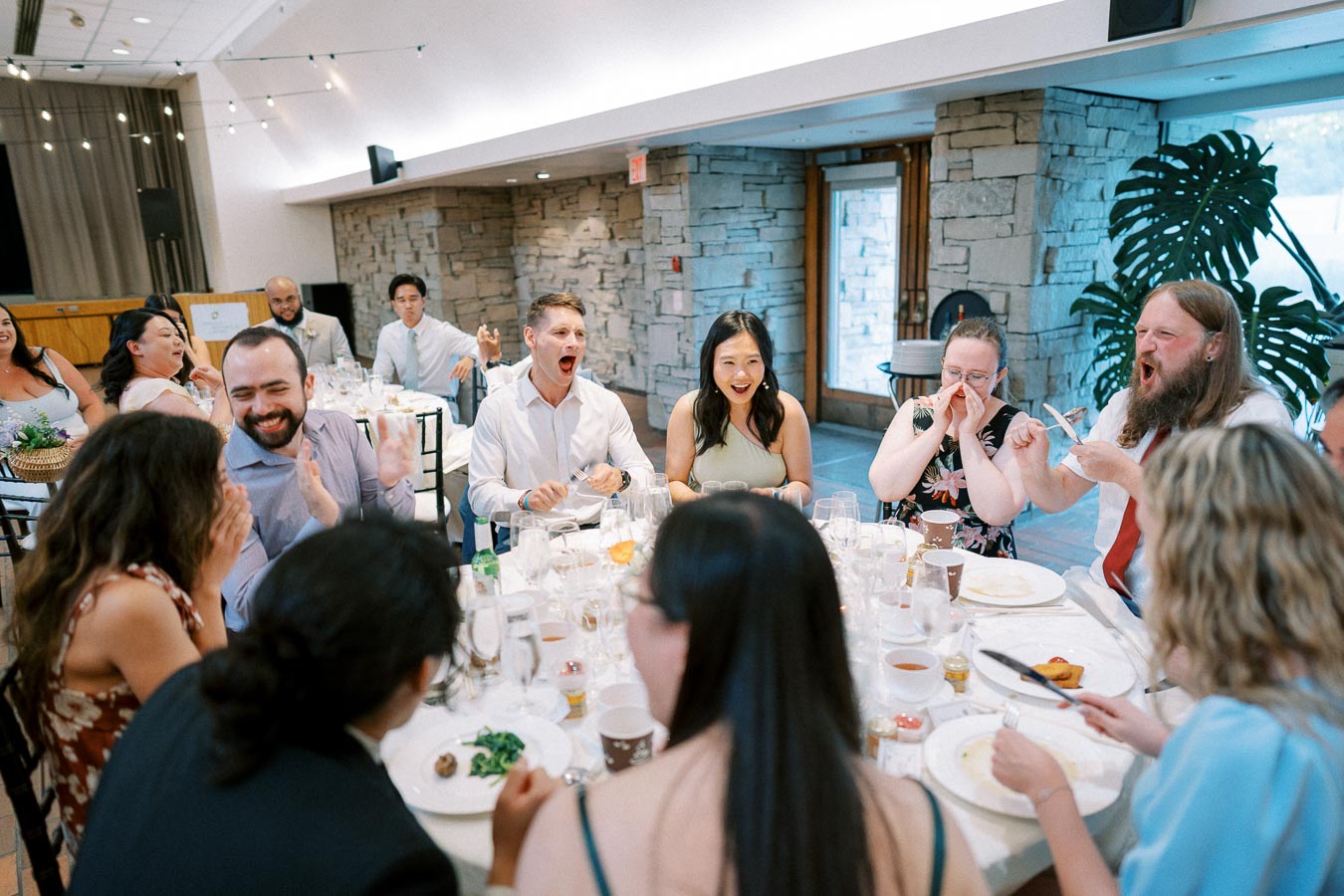A lively group of people enjoying a meal together at a wedding reception, seated around a beautifully set round table indoors, with visible laughter and conversation adding to the joyful atmosphere.