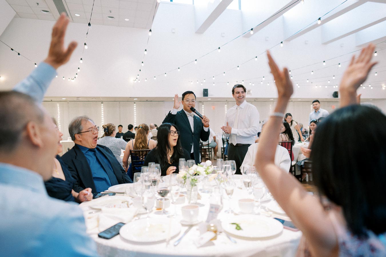 A lively indoor wedding reception with guests seated around a round table, hands raised in excitement, as a speaker wearing a suit and holding a microphone engages with the audience. The venue is adorned with string lights, creating a festive atmosphere.