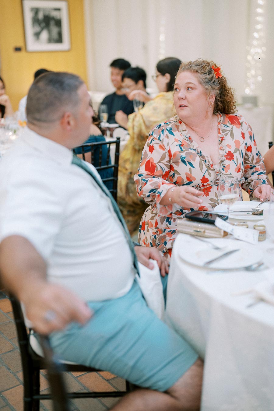 A woman in a floral dress and a man in a white shirt with blue shorts engage in conversation at a formal event, seated at a table set with dinnerware and glasses, surrounded by other attendees.