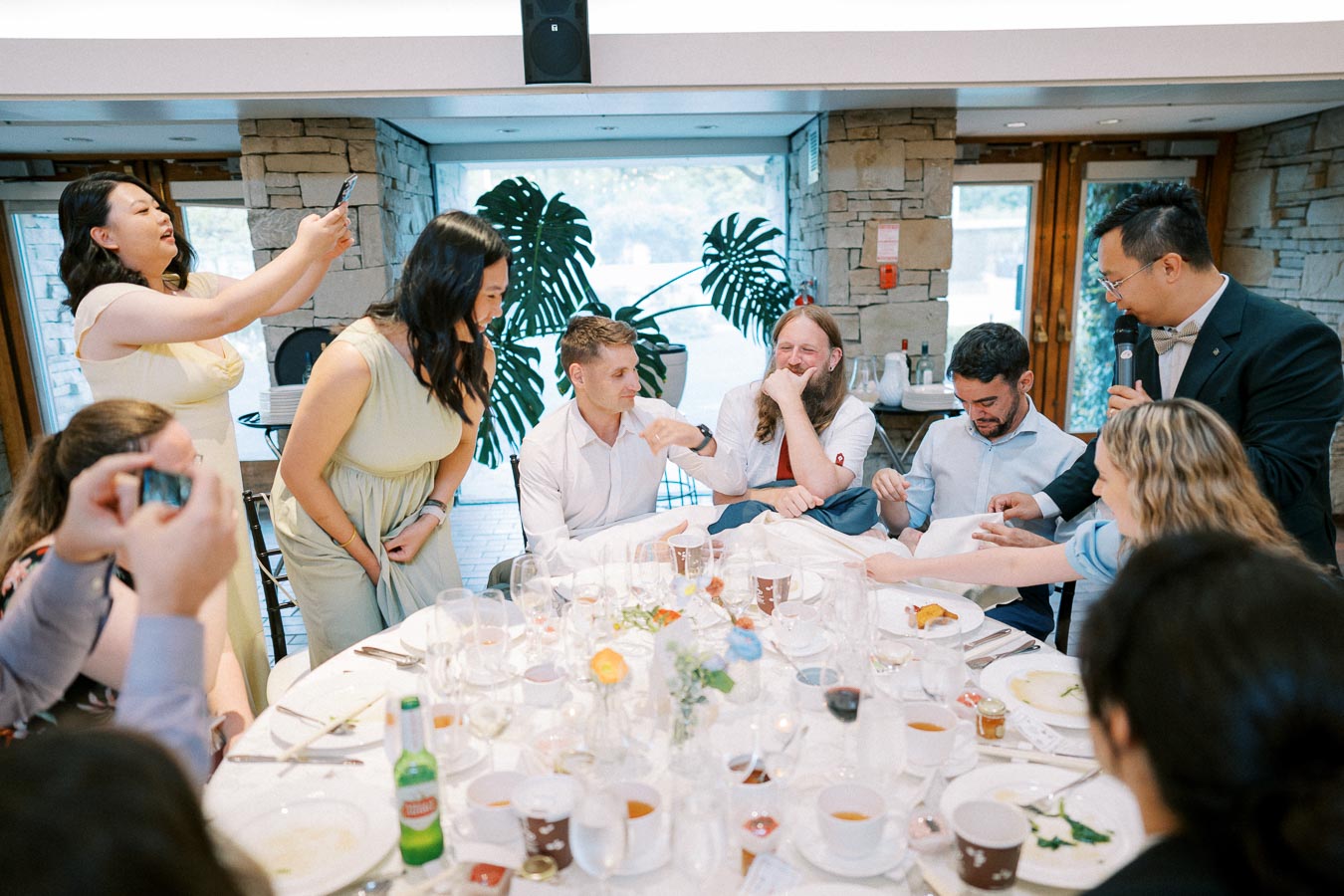A lively gathering of people enjoying a celebration at a banquet table in a well-lit indoor venue, with one person taking photos and another speaking with a microphone.