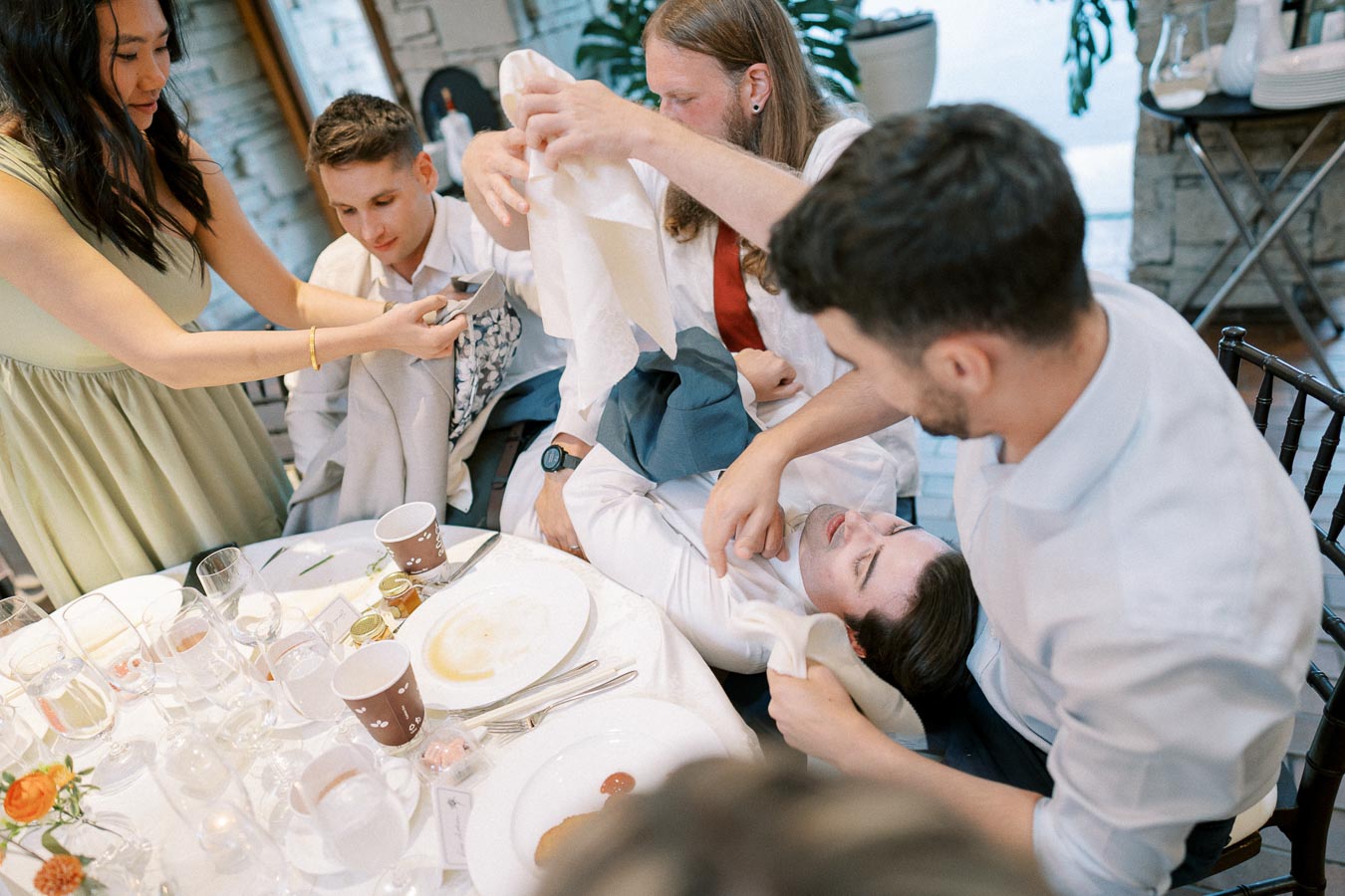 A group of people playfully interacting around a dining table, with dishes and drinks, in a lively social gathering setting.