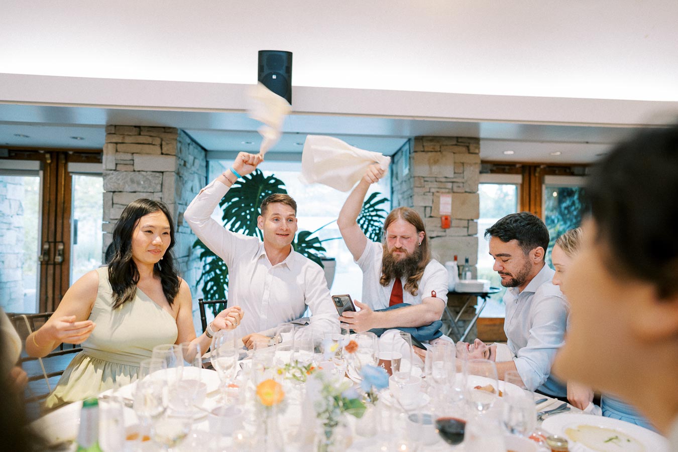 A group of people enjoying a celebration at a dining table in a rustic setting, with one person enthusiastically waving a cloth napkin in the air. The table is set with glassware, flowers, and candles, creating a festive atmosphere.