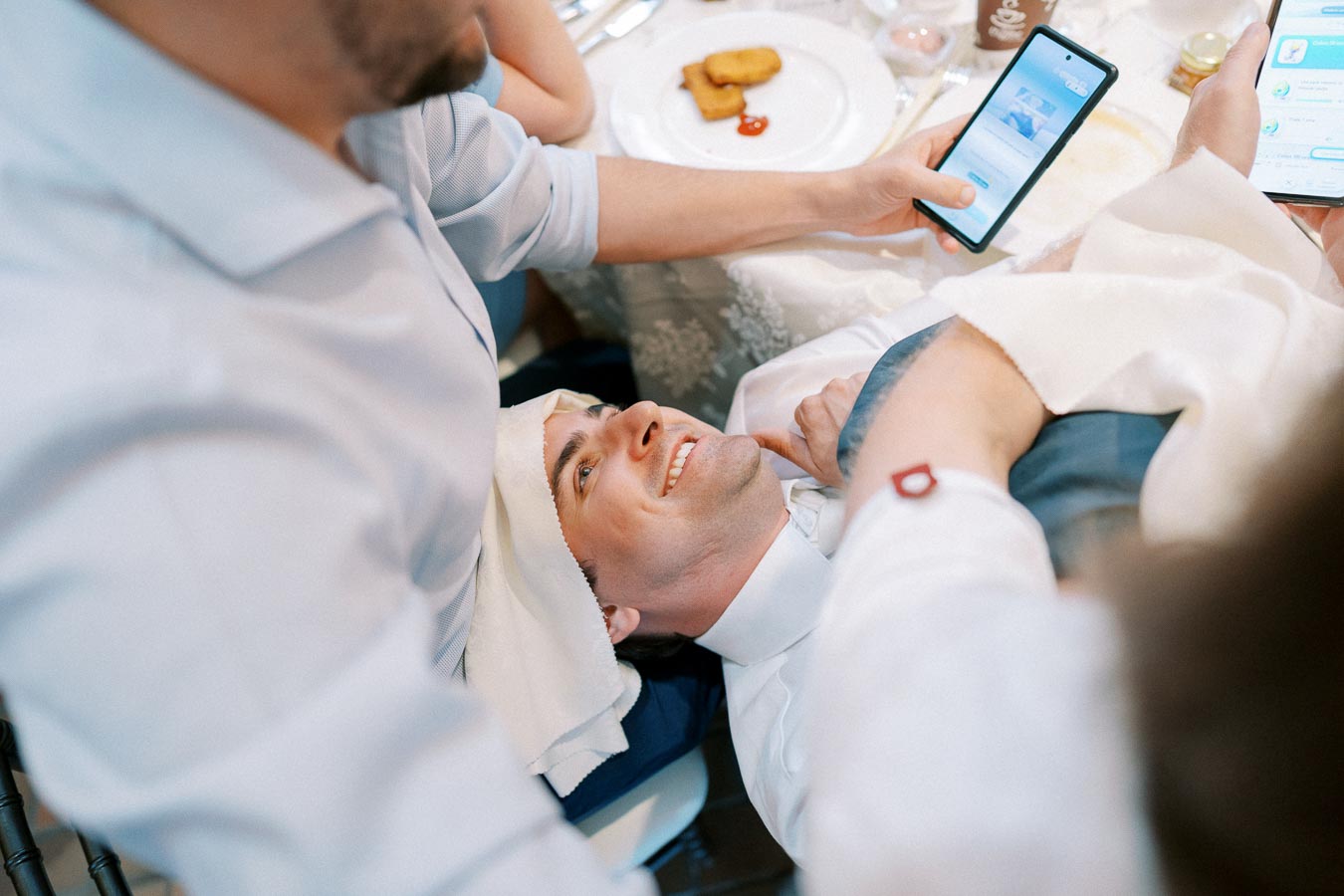A person lying on a chair at a table, smiling, while others interact with their smartphones, with food visible on the table, suggesting a casual social gathering.