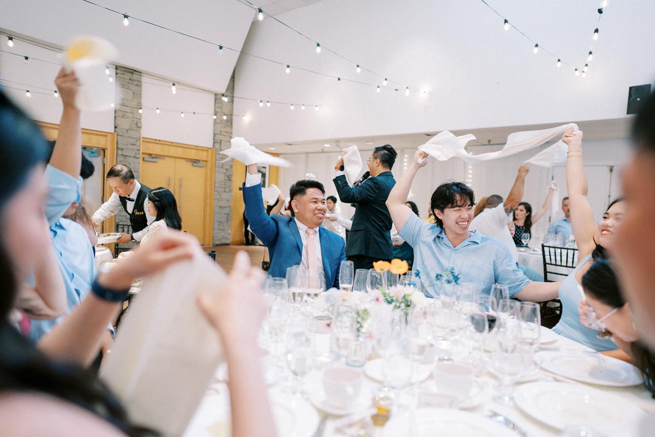 Guests joyfully waving napkins at an indoor wedding reception, with a festive atmosphere and string lights overhead.