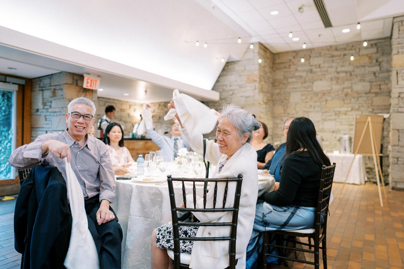 Guests joyfully waving napkins at a wedding reception, seated around a decorated table in a warmly-lit venue with stone walls.