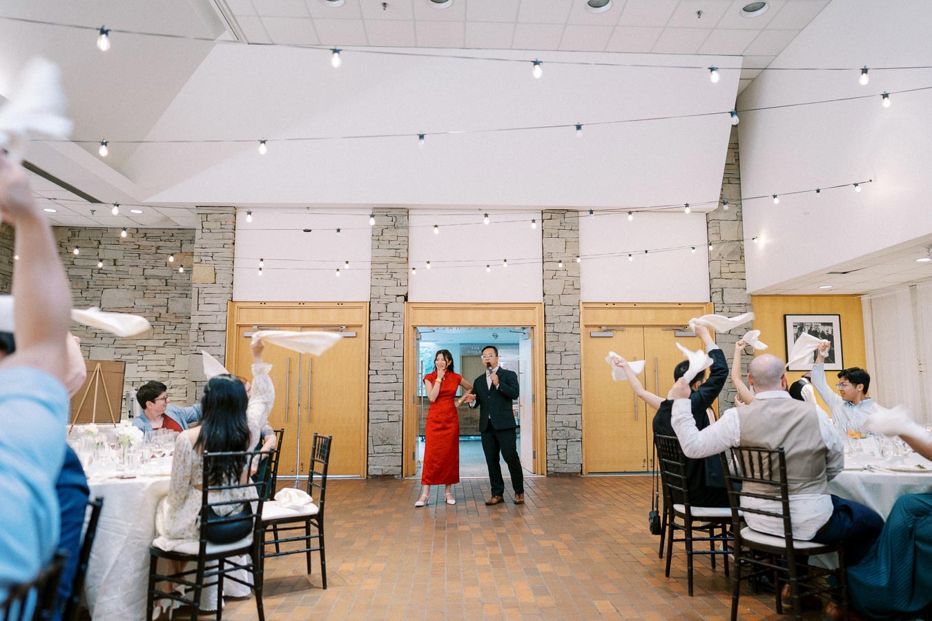 Guests enthusiastically wave napkins at a wedding reception while the couple addresses the crowd, with a backdrop of string lights and stone walls.