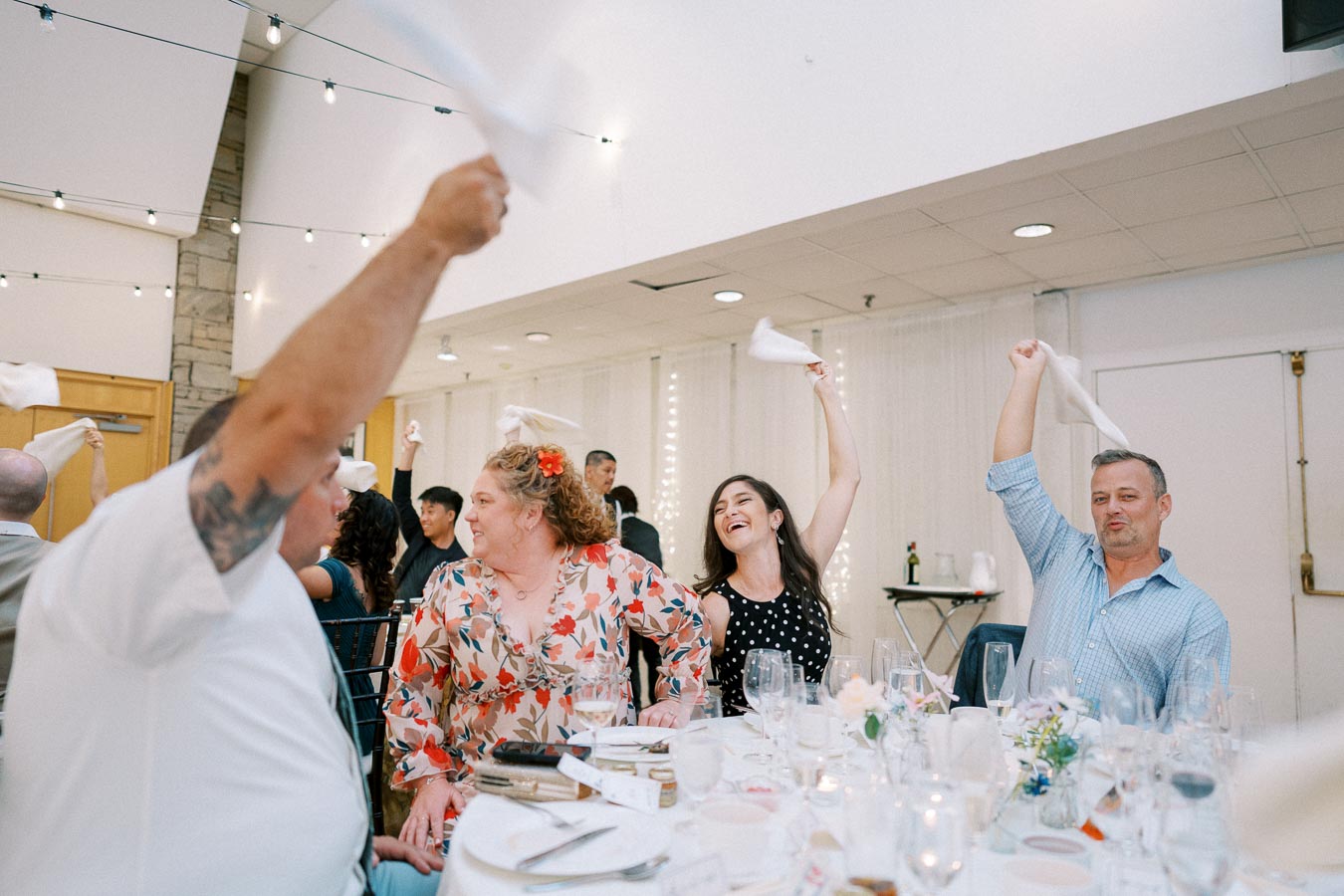 A joyful group of people waving napkins in the air at a festive indoor celebration, surrounded by dining tables, lights, and decorations.