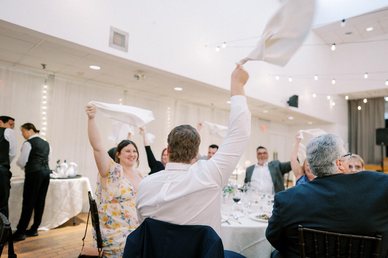 Guests joyfully wave napkins in celebration at a wedding reception, seated around a decorated table in a warmly lit venue.