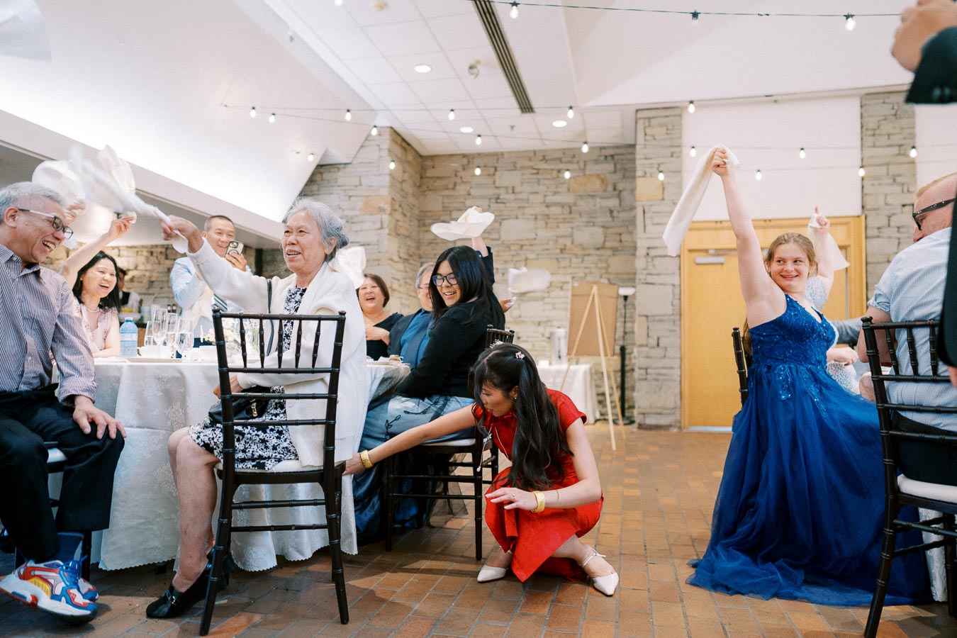 Guests enjoy a lively celebration at a joyful event, waving napkins and smiling around a decorated table, capturing the essence of fun and togetherness in a beautifully lit venue.
