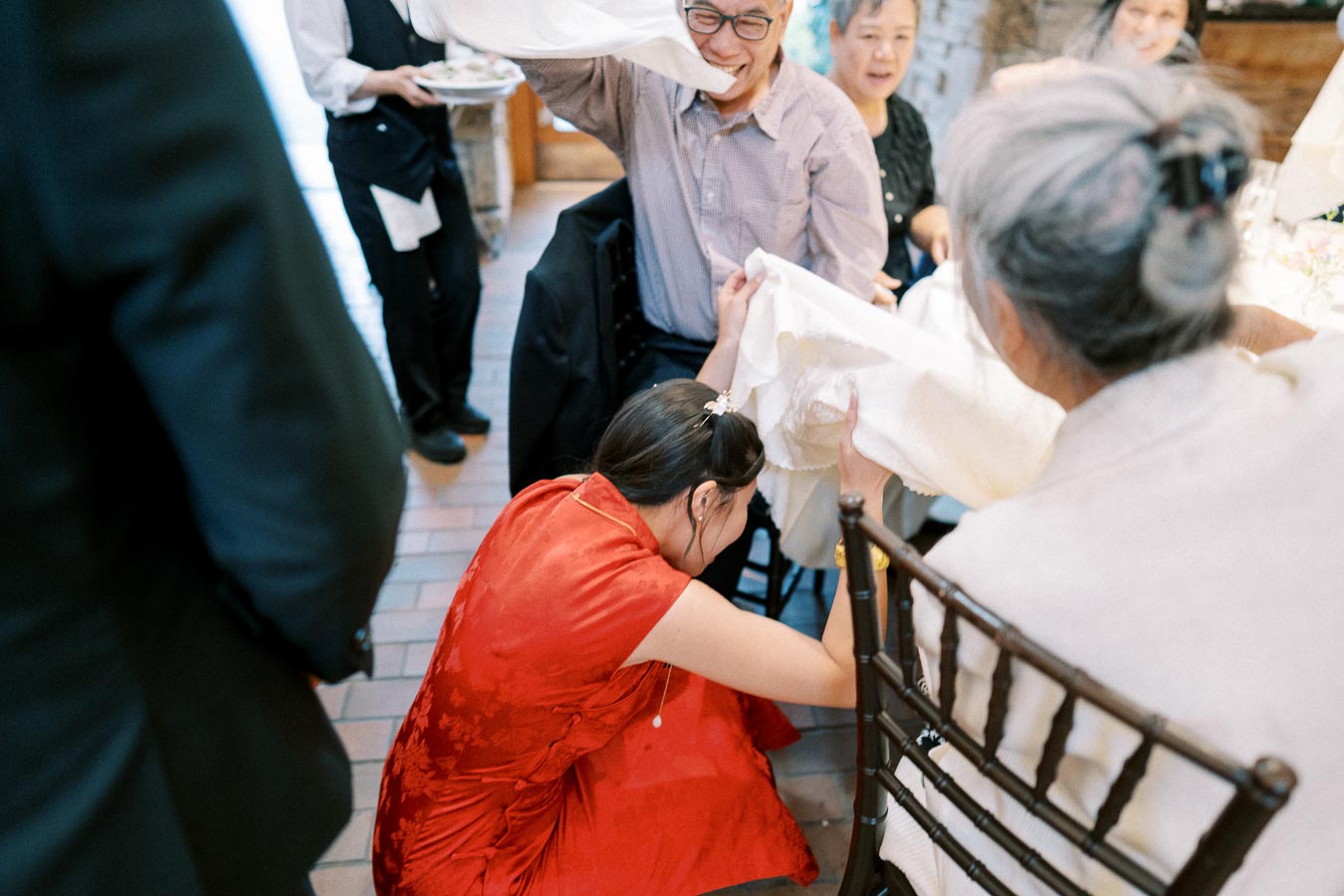 A person in a red dress crouches with family members at a dining table, participating in a joyful cultural ceremony, with laughter and conversation all around.