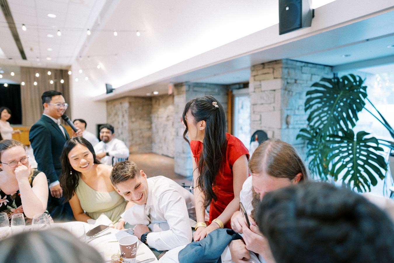 A group of people enjoying a lively celebration at an indoor event, featuring guests smiling and interacting around a table adorned with wine glasses and a palm plant in the background.