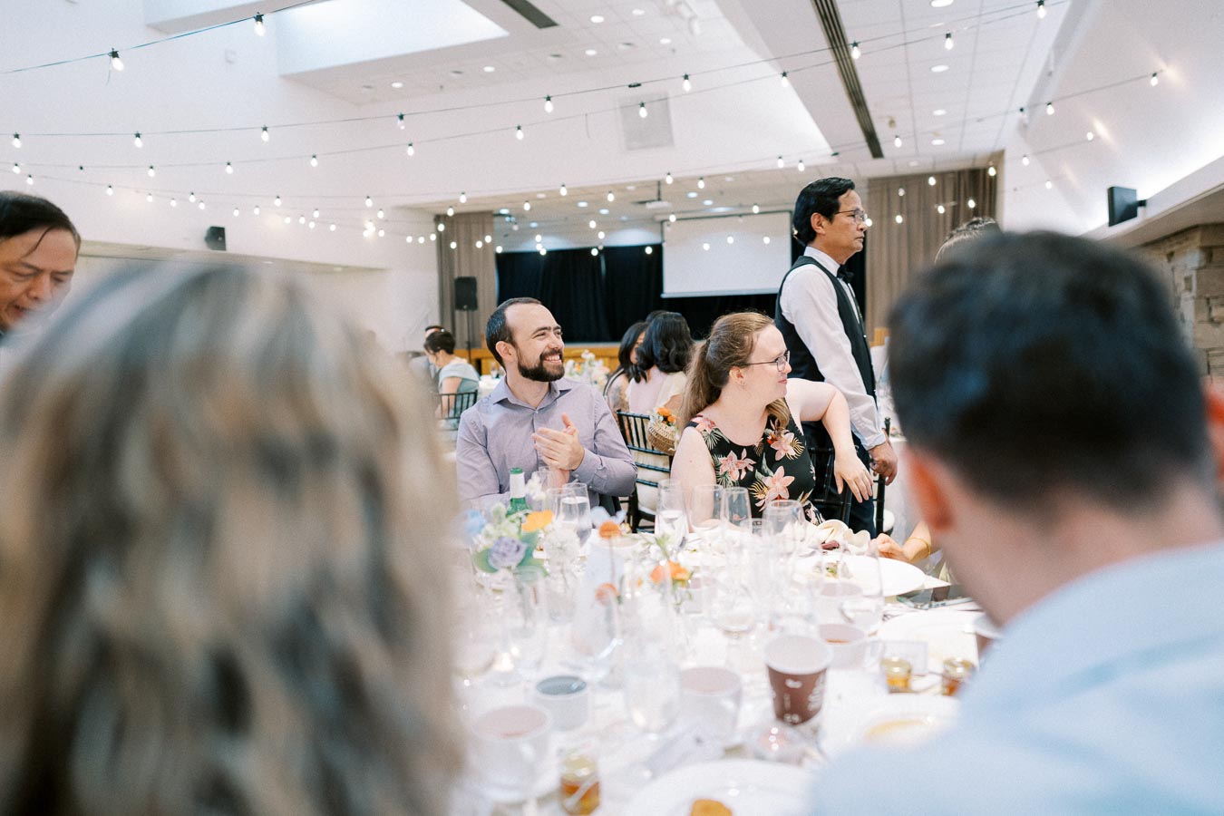 A group of people smiling and engaged in conversation at a beautifully decorated indoor event with string lights overhead, surrounded by elegantly set tables with flowers and glassware.