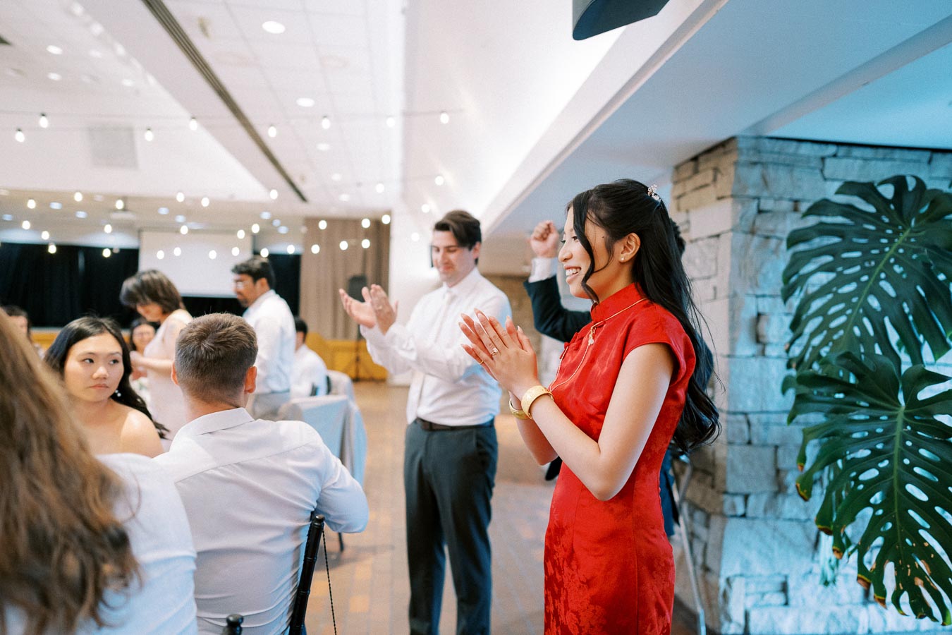 A woman in a red traditional dress clapping joyfully at an indoor event, surrounded by guests in a well-lit space decorated with string lights.