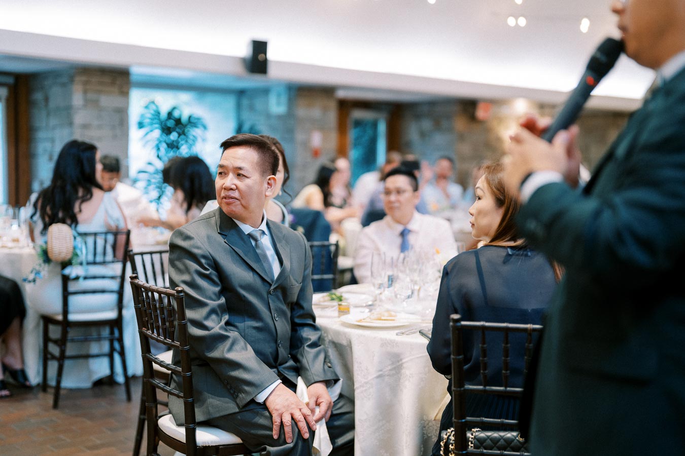 Guests seated at a formal event, with a man in a suit looking to the side and tables set with white tablecloths in the background, suggesting a sophisticated wedding or banquet atmosphere.