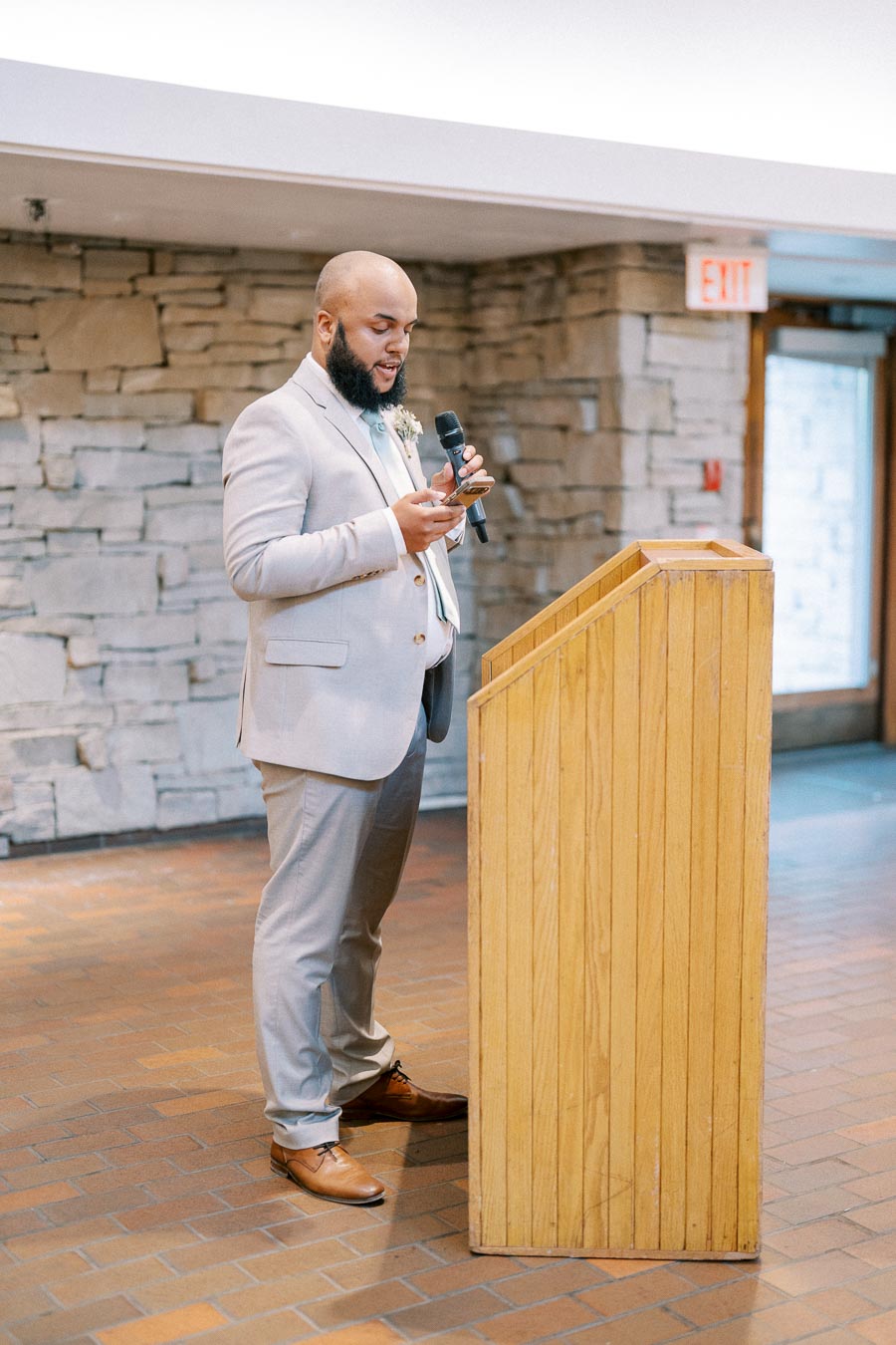 A man in a light gray suit giving a speech at a podium indoors, holding a microphone and phone, with a stone wall background.